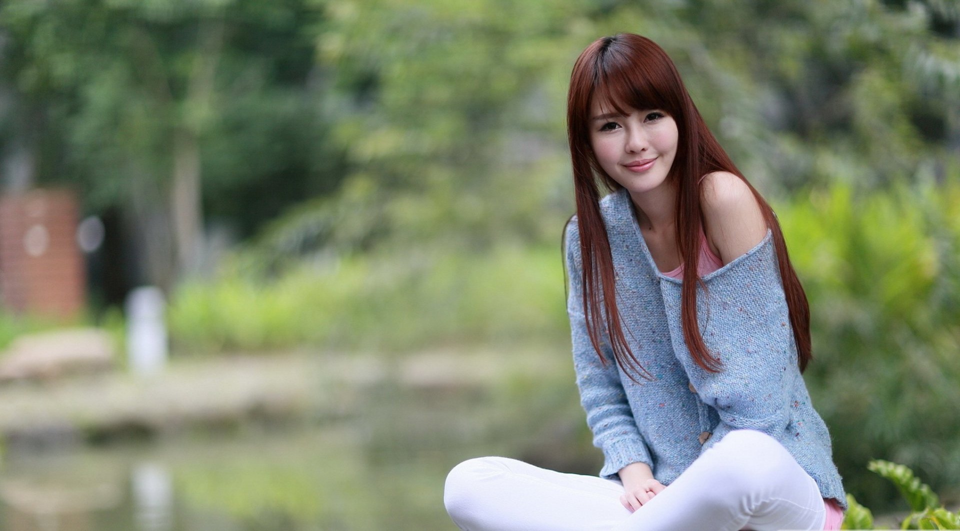 HD desktop wallpaper featuring an Asian woman with long brown hair, wearing a blue sweater and white pants, sitting outdoors near a calm, green natural setting.