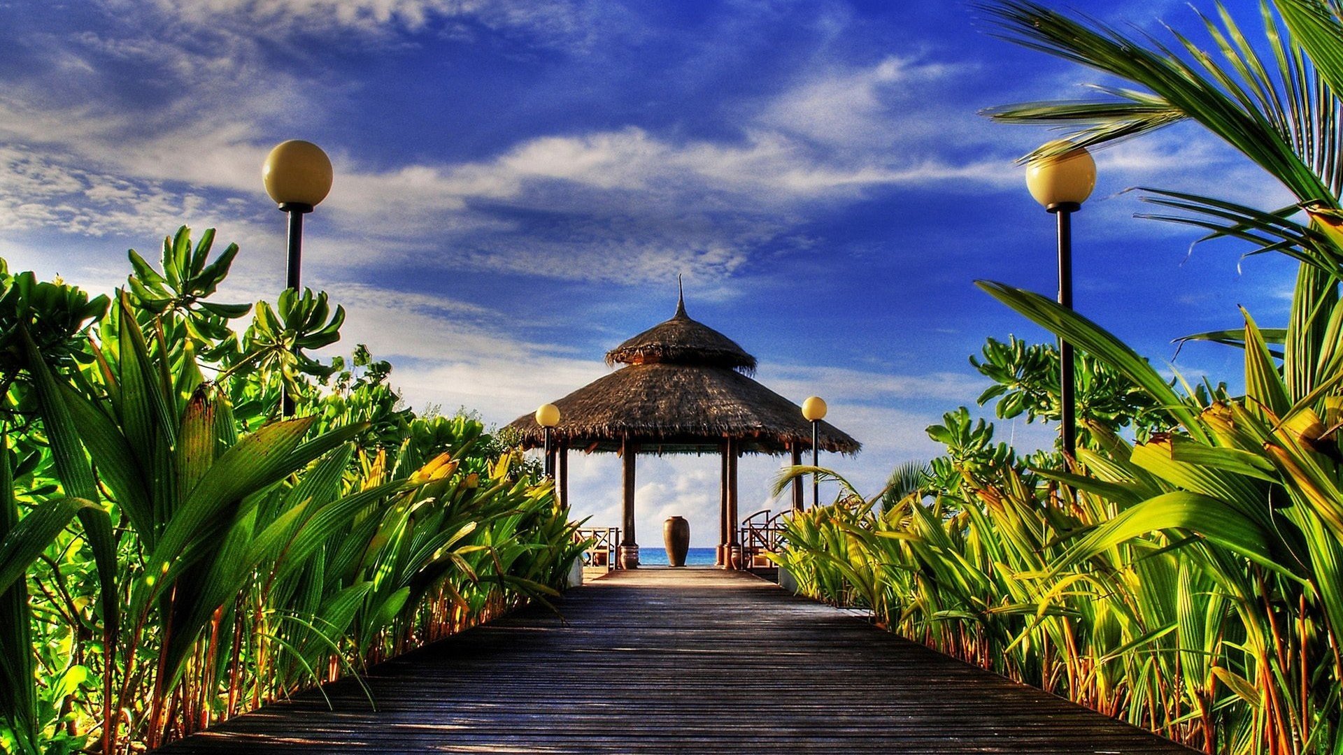 HD PC desktop wallpaper featuring a vibrant tropical scene with lush greenery, a wooden walkway, and a thatched gazebo under a partly cloudy blue sky.