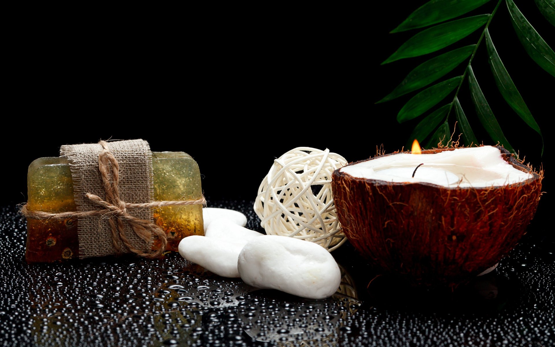 HD desktop wallpaper featuring a man-made spa setting with soap bars, a lit candle in a coconut shell, decorative spheres, and green leaves on a reflective black surface.