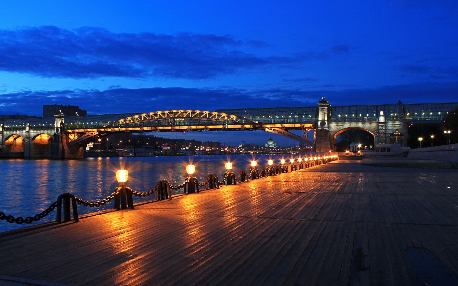 A stunning HD desktop wallpaper featuring a beautifully lit bridge at dusk, reflecting on the water, surrounded by a warm glow from lamps along the walkway.