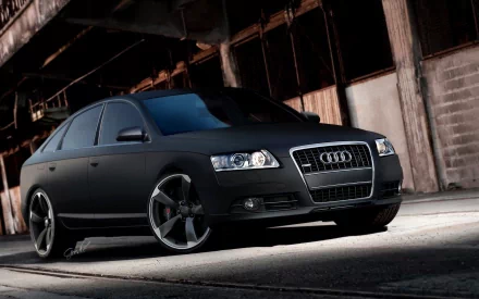 A sleek black Audi A6 parked in an industrial setting, showcasing its modern design and impressive wheels, set against a dramatic backdrop. HD desktop wallpaper and background image.