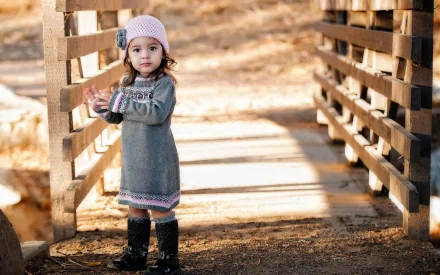 HD desktop wallpaper featuring a young child with a pink hat and gray dress standing on a wooden bridge. The sunlight casts long shadows, highlighting the serene outdoor setting.