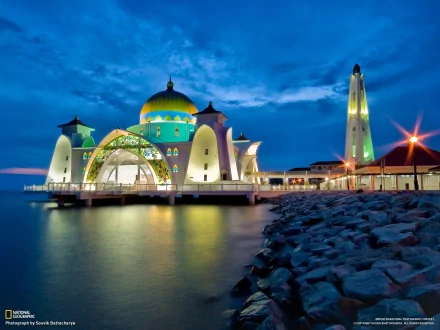 HD desktop wallpaper showcasing the Malacca Straits Mosque at dusk, beautifully illuminated against a deep blue sky with calm waters and rocky shoreline.