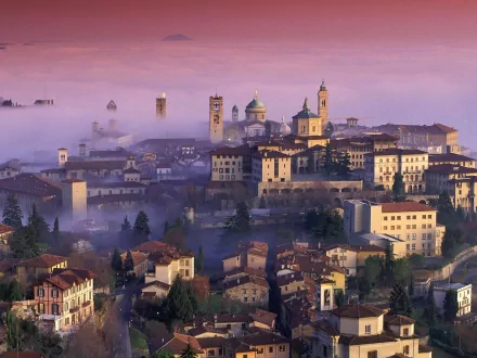 A stunning HD image of Bergamo, Italy, showcasing a picturesque town enveloped in morning mist, with historic buildings and clock towers rising above the fog.