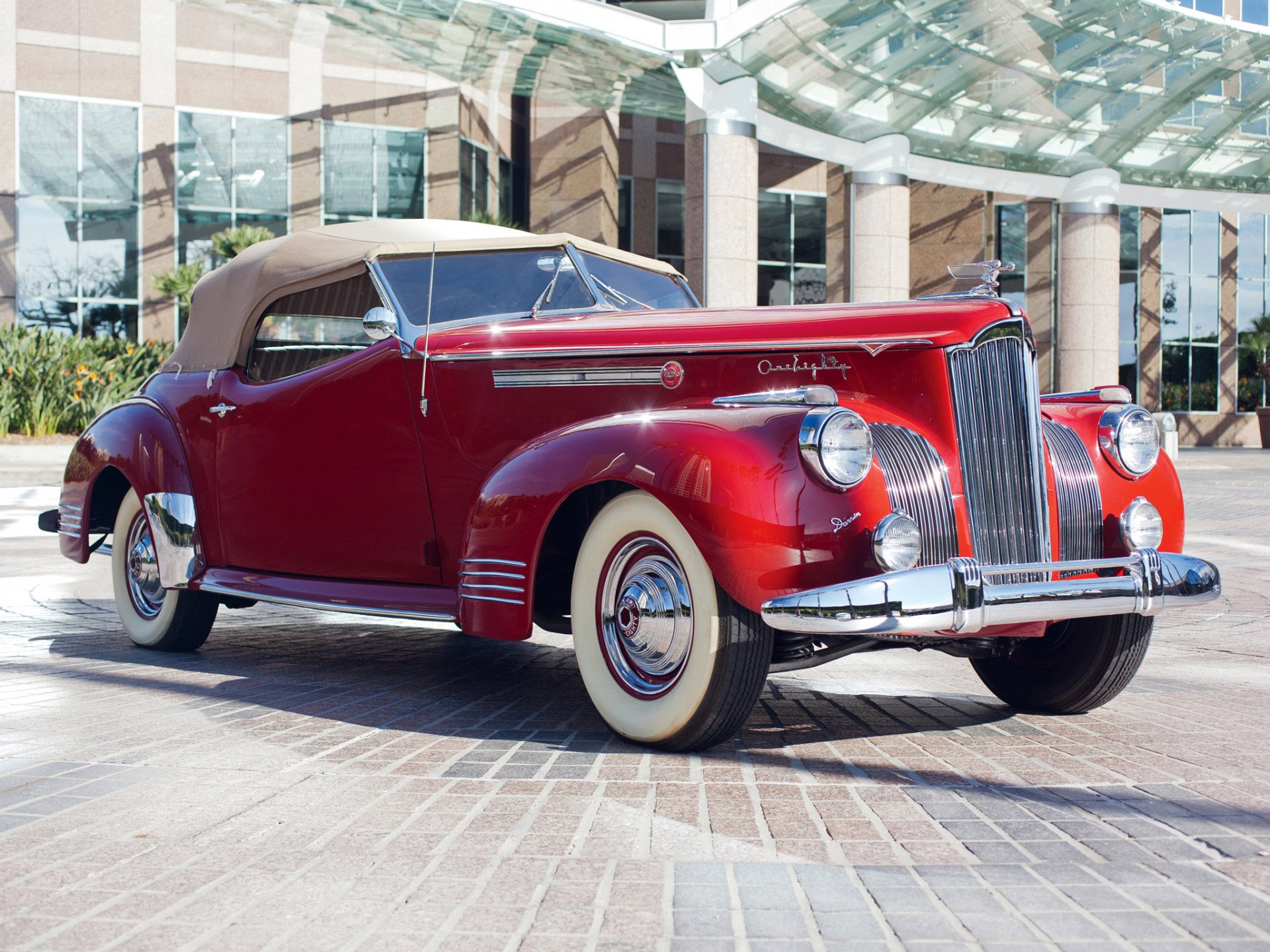 A classic red Packard 180 convertible parked in front of modern glass buildings, displayed as a high-definition PC desktop wallpaper and background.