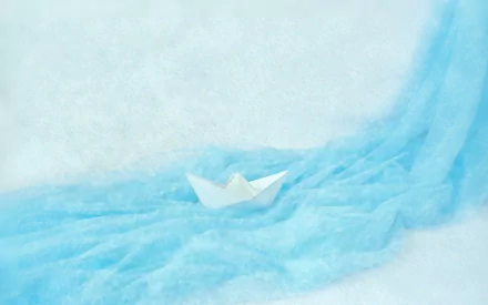 2K Quad HD PC desktop wallpaper showing a white origami paper boat (man-made) drifting on soft blue fabric waves.