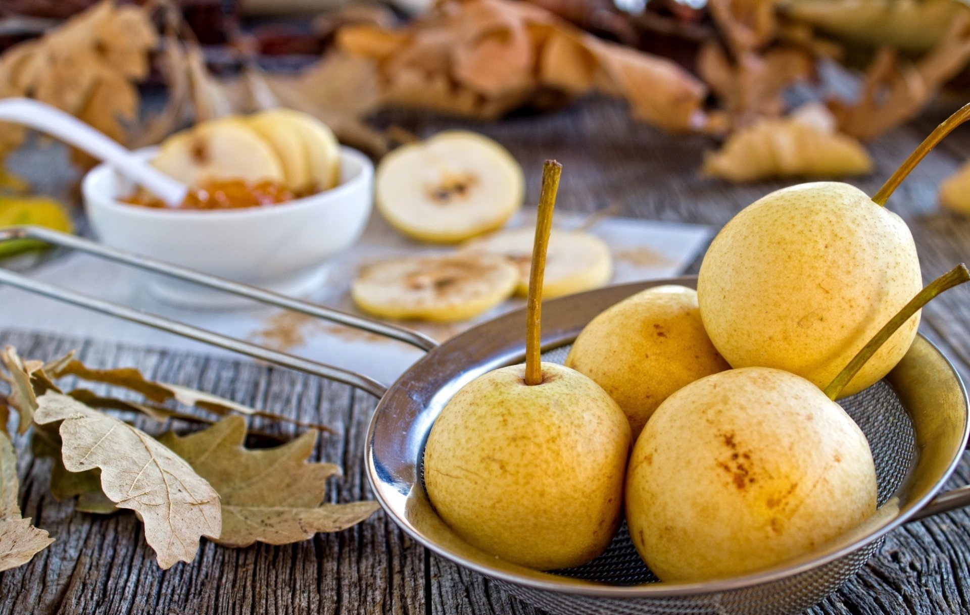 HD desktop wallpaper featuring a close-up of fresh pears in a metal bowl on a rustic wooden surface with dried leaves and sliced pears in the background.