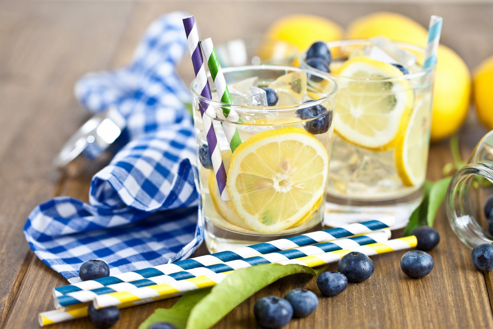 HD desktop wallpaper featuring refreshing cocktails with lemon slices, blueberries, and colorful striped straws on a wooden surface with a blue checkered cloth.