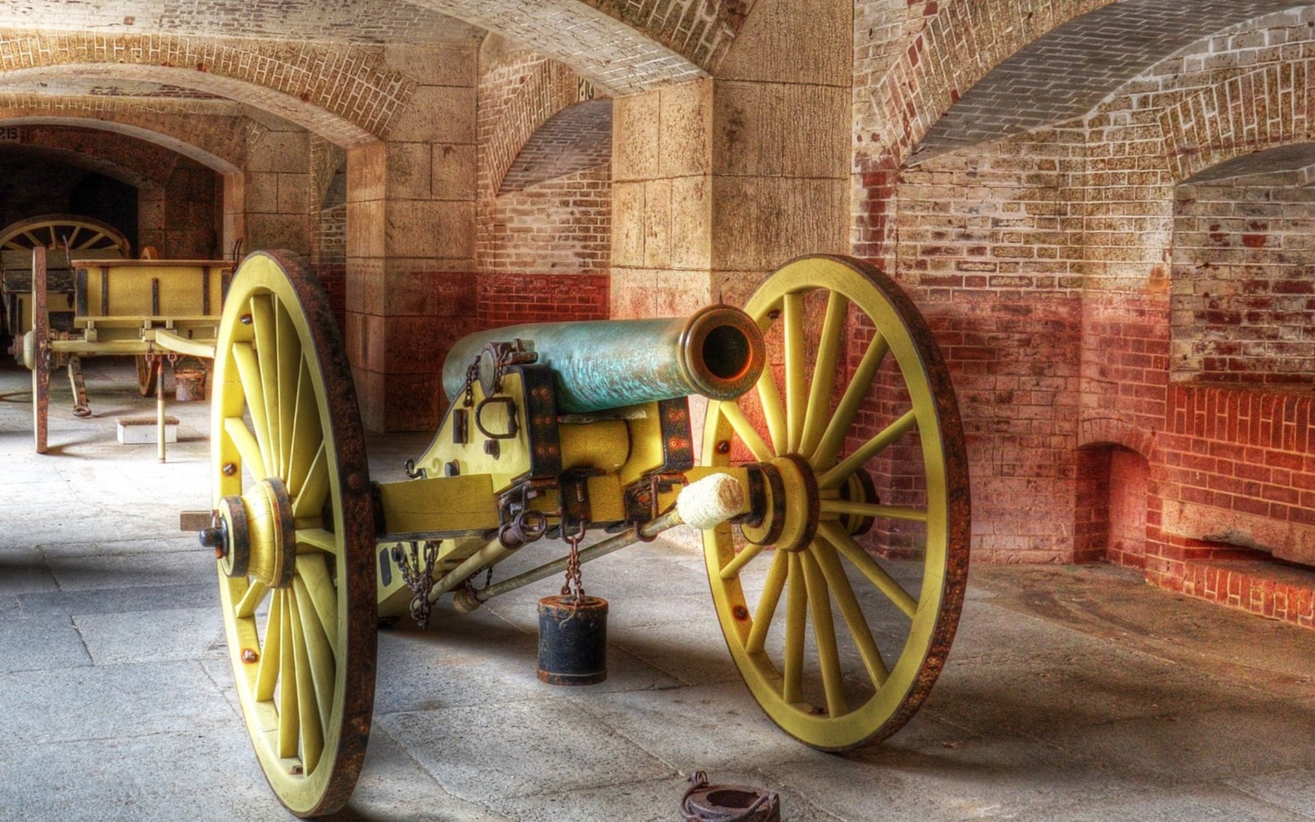 HD PC desktop wallpaper of a historic military artillery cannon with large yellow wheels inside a brick-arched fort or storage room.