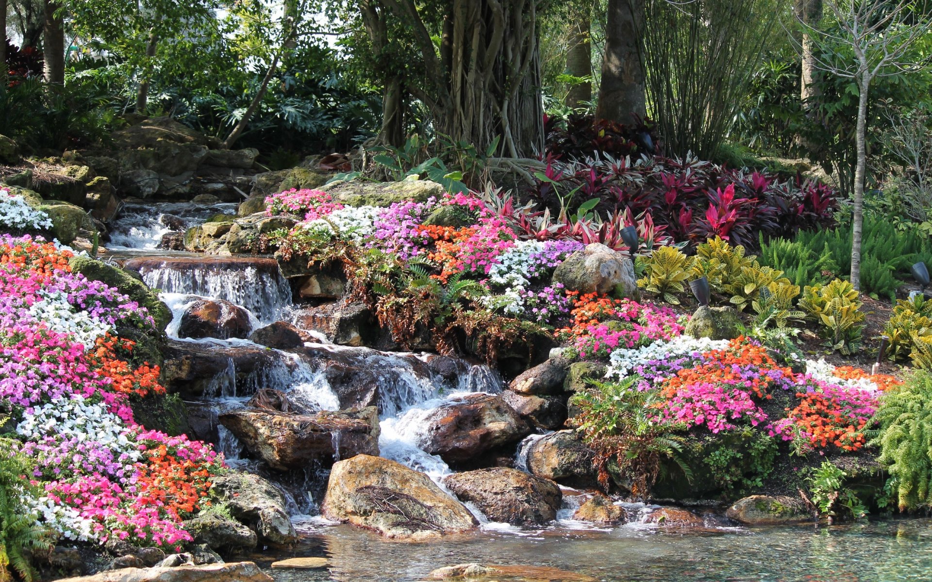 A vibrant waterfall cascades over rocks, surrounded by colorful flowers and lush greenery, creating a serene nature scene for a stunning HD PC desktop wallpaper.
