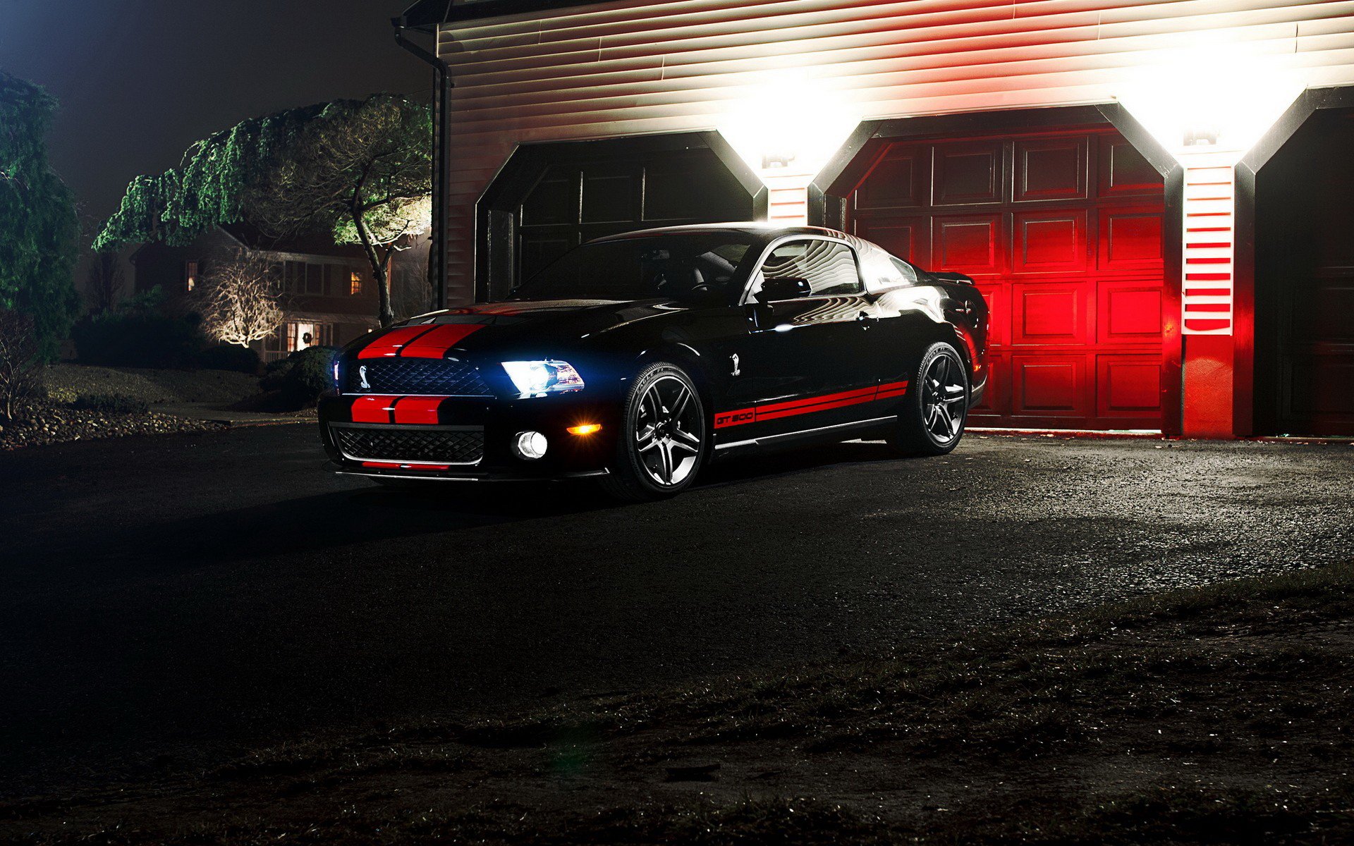 A sleek Ford Mustang Shelby GT500 parked in front of a garage at night, illuminated by soft red and white lights, creating a striking HD desktop wallpaper and background.