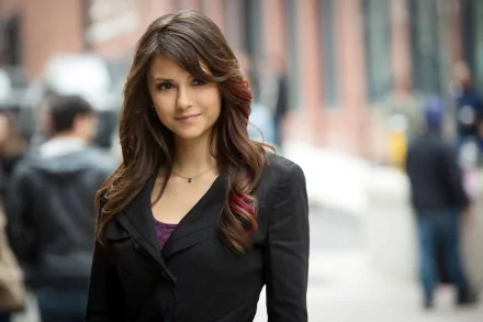 A confident Nina Dobrev poses outdoors, showcasing her stylish black coat and flowing hair, set against a softly blurred city background. A striking HD wallpaper for fans.