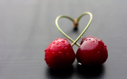 HD desktop wallpaper featuring two red cherries with water droplets, their stems intertwined to form a heart shape on a dark background.