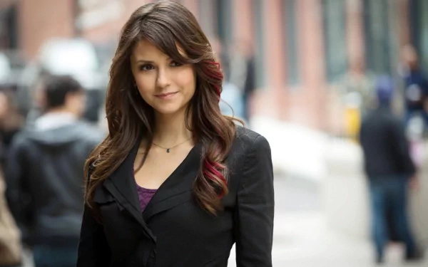 A confident Nina Dobrev poses outdoors, showcasing her stylish black coat and flowing hair, set against a softly blurred city background. A striking HD wallpaper for fans.
