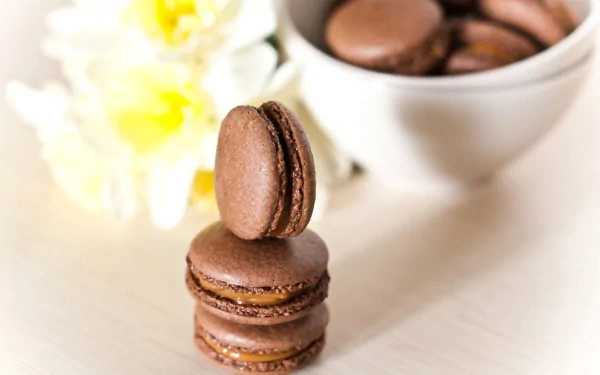 A stack of chocolate macarons sits in front of a bowl filled with more, accompanied by delicate flowers, creating an enticing dessert wallpaper for your desktop.