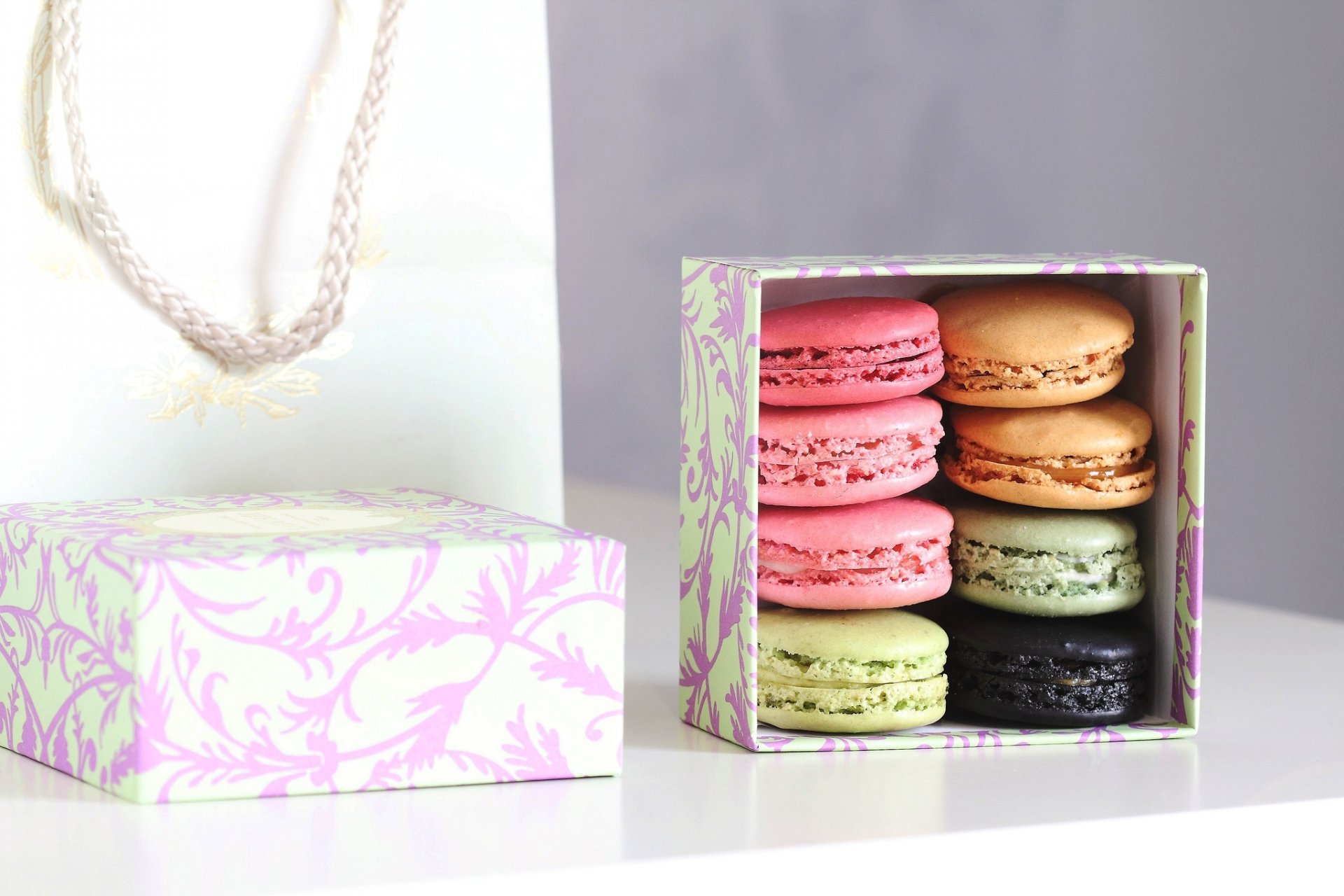 HD PC desktop wallpaper featuring a pastel-colored box of assorted macarons with pink, beige, green, and black hues, alongside a matching gift bag on a white surface.