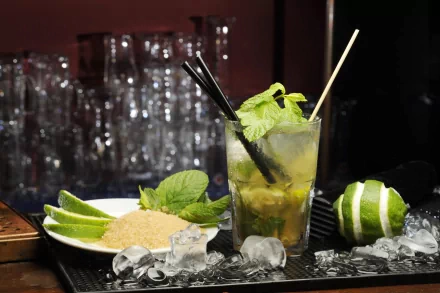 A refreshing cocktail featuring mint and lime, served over ice, alongside a plate of brown sugar and lime wedges, set against a backdrop of glassware. 4K Ultra HD desktop wallpaper.