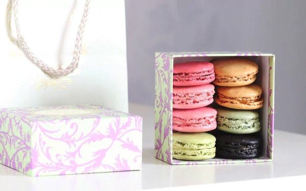 HD PC desktop wallpaper featuring a pastel-colored box of assorted macarons with pink, beige, green, and black hues, alongside a matching gift bag on a white surface.