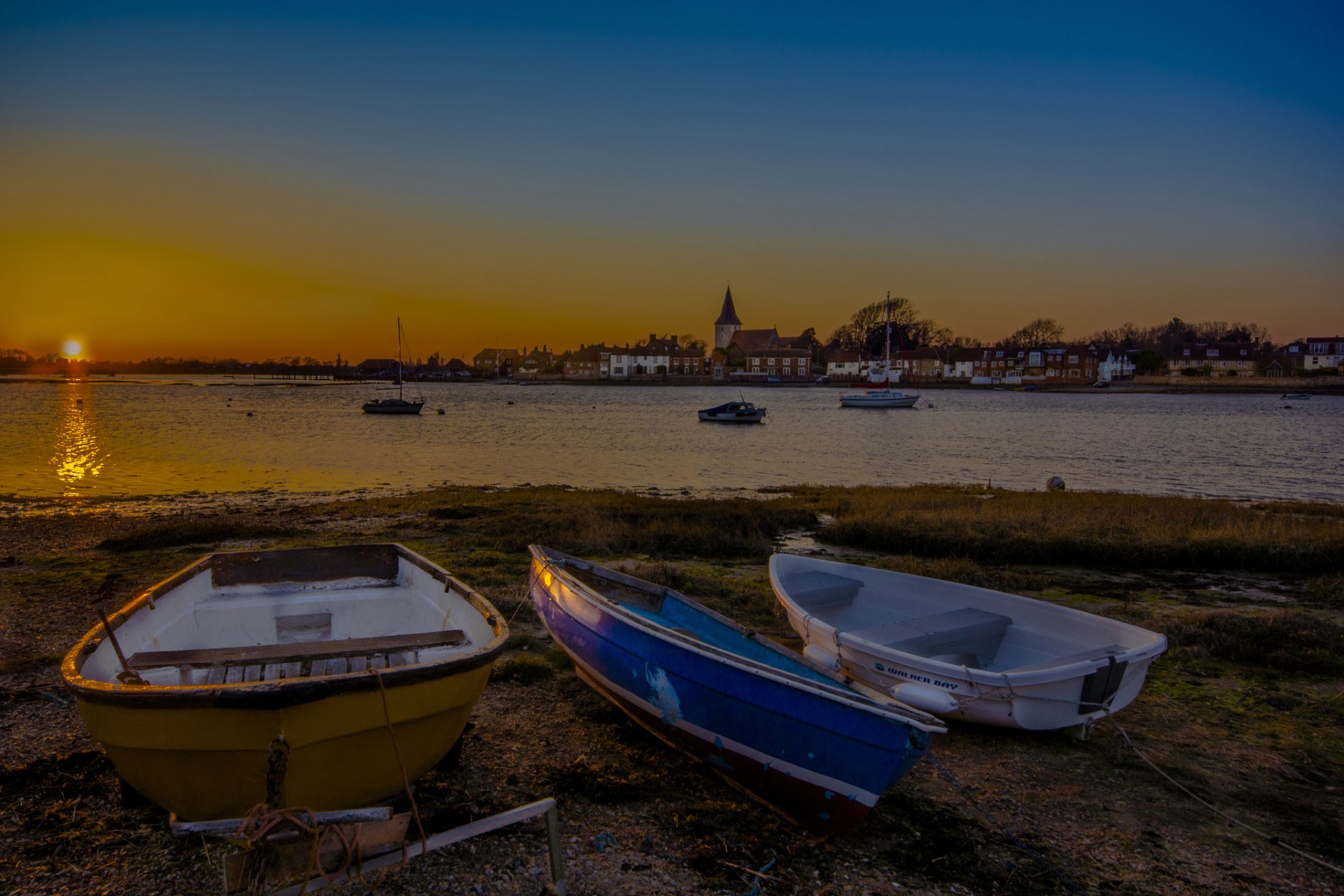 HD desktop wallpaper featuring three small boats resting on a shore at sunset, with calm water and a distant town under a gradient sky.