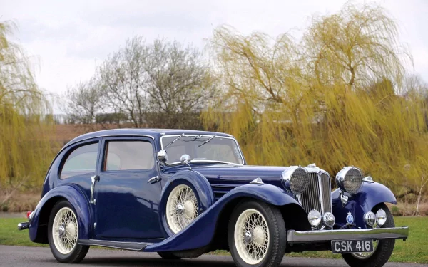 A classic Jaguar SS100 vintage vehicle in dark blue, set outdoors with trees and cloudy sky, featured as an HD PC desktop wallpaper and background.