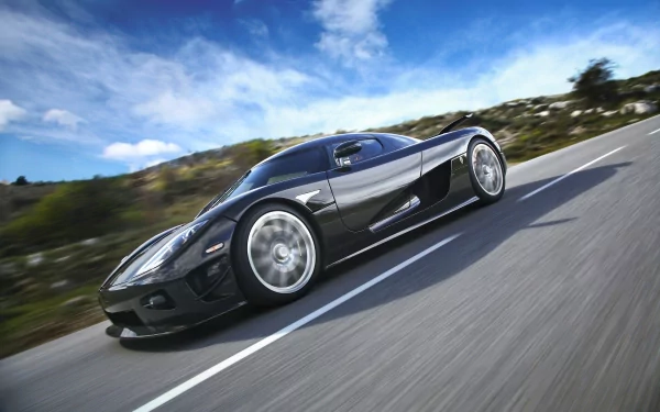 A sleek black Koenigsegg CCX speeds down a scenic road, showcasing its aerodynamic design against a backdrop of blue skies and rolling hills. High-definition wallpaper for any desktop.