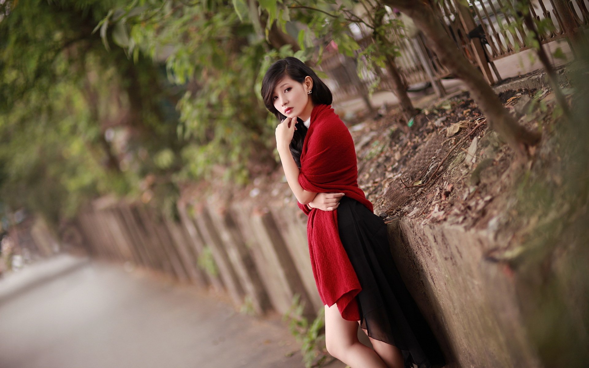 A woman in a red and black outfit poses thoughtfully against a scenic background, surrounded by greenery. This HD image serves as an elegant desktop wallpaper.