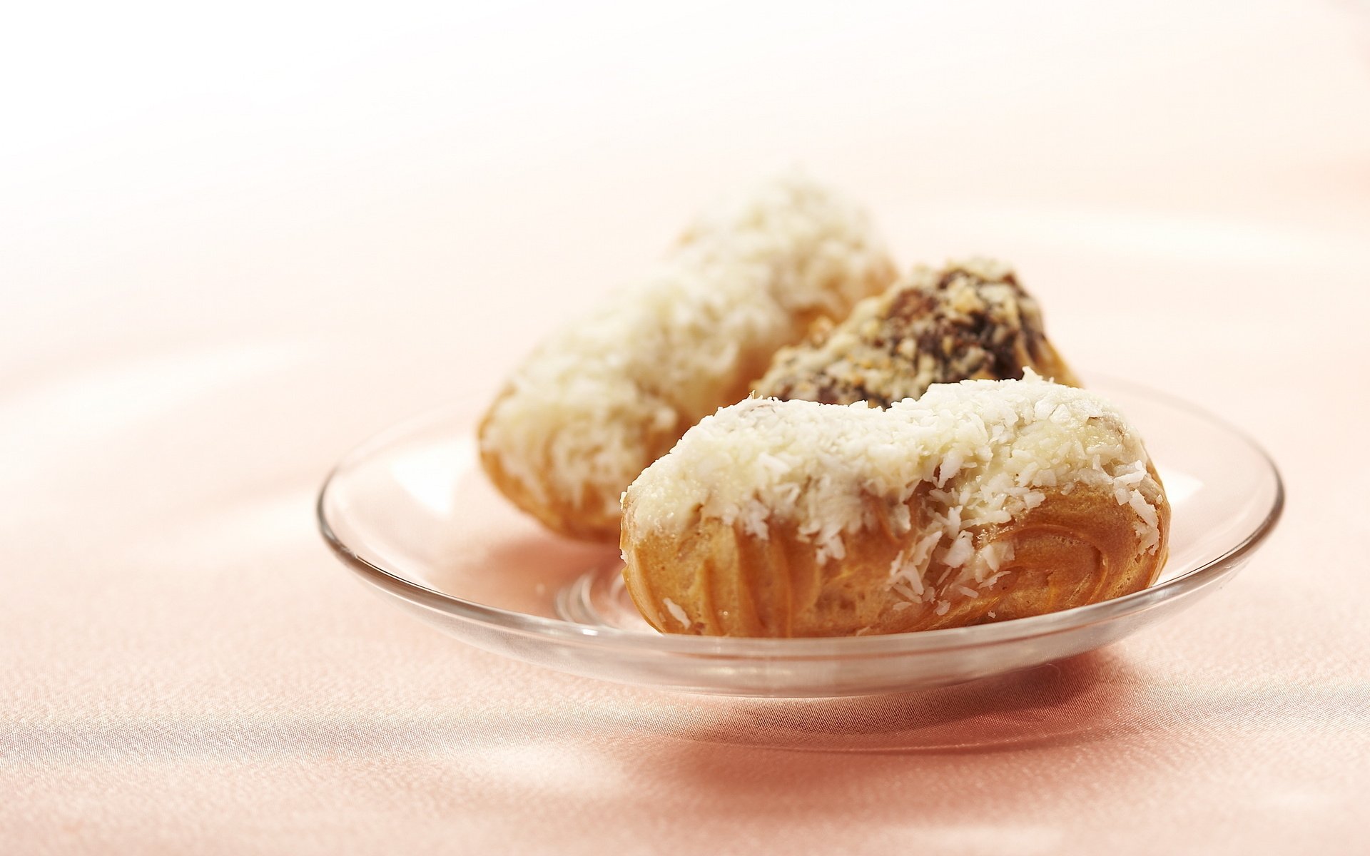 HD desktop wallpaper featuring a clear glass plate with three assorted sweets coated in white and chocolate toppings on a soft pink background.
