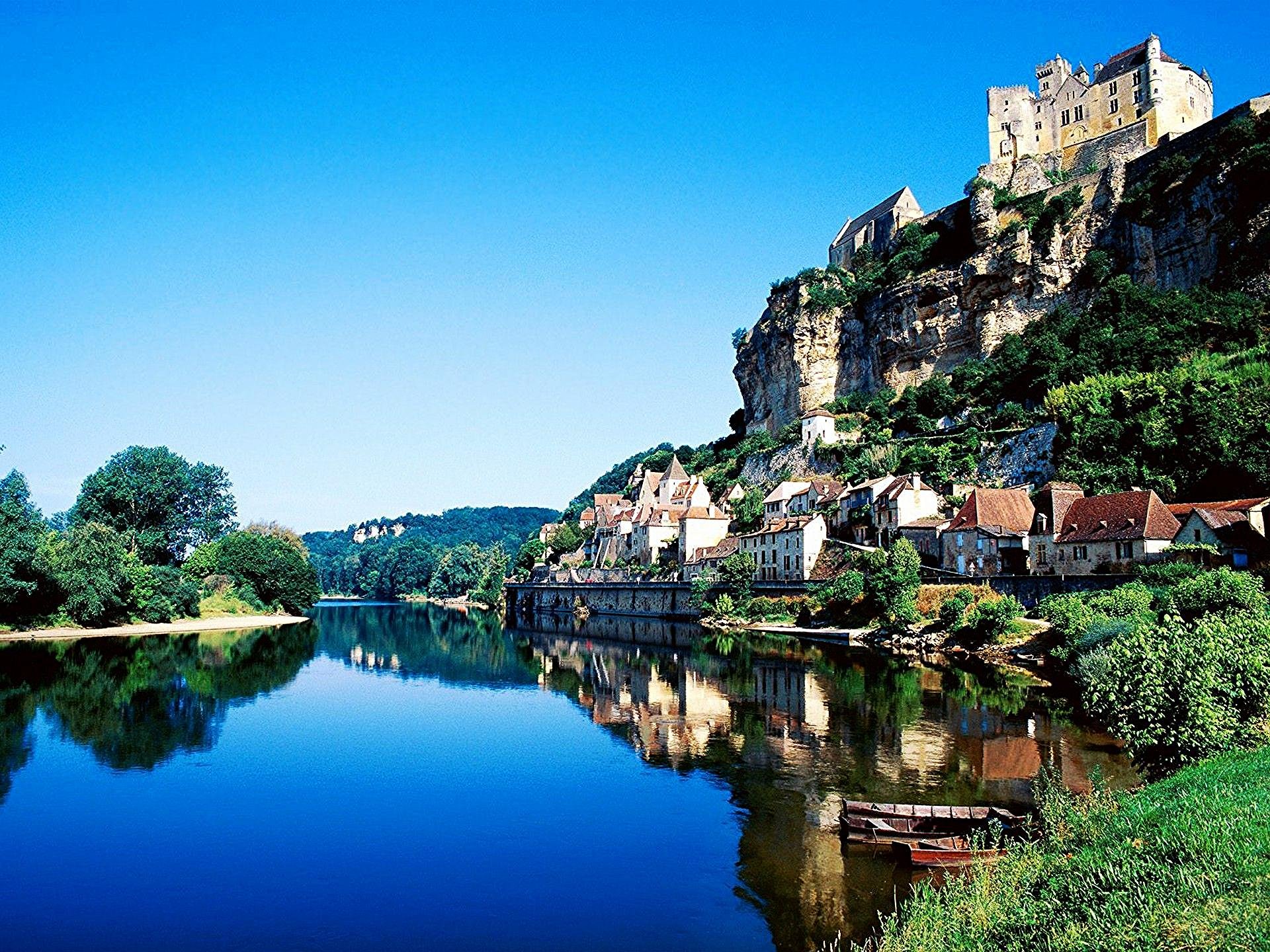 A serene landscape featuring a clear blue sky, a calm river reflecting a quaint village and a castle perched on a rocky hill, creating a stunning HD desktop wallpaper.
