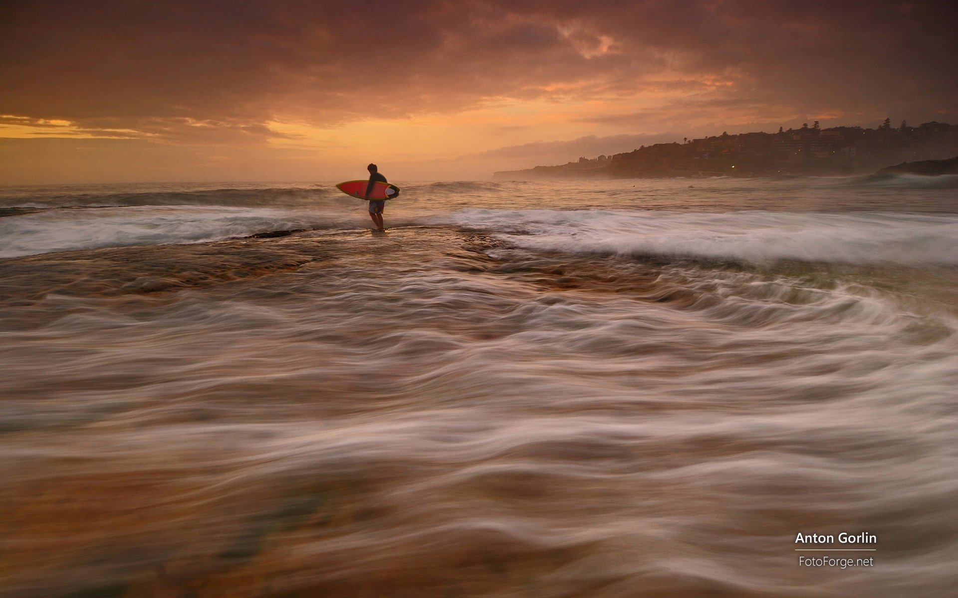 HD PC desktop wallpaper/background: lone surfer carrying a board into churning surf at sunset — surfing, sports scene with dramatic sky and motion-blurred waves.