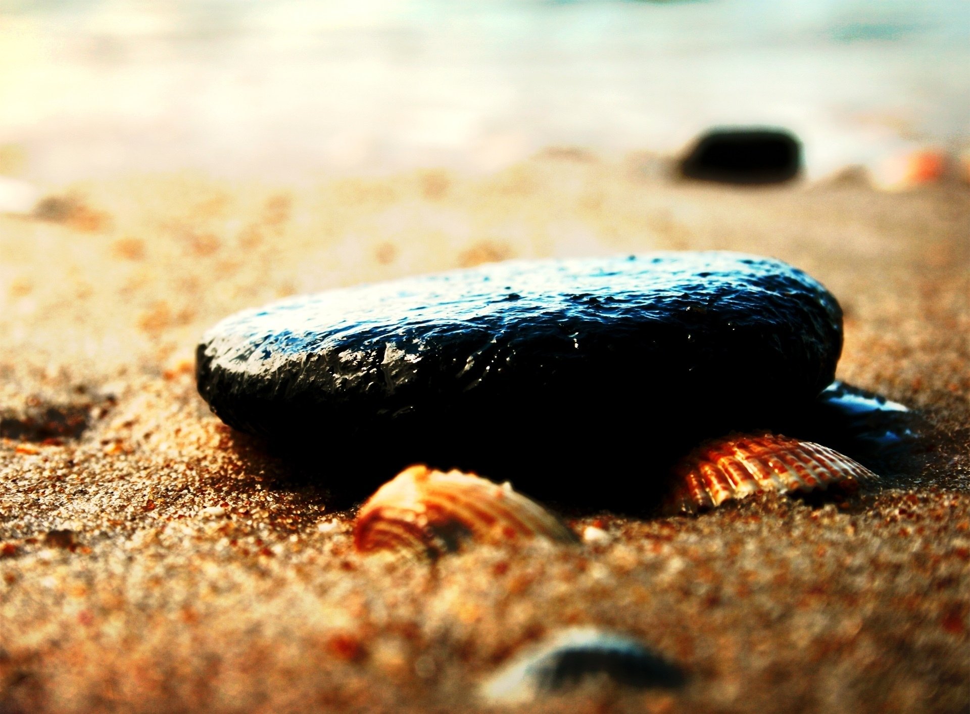 Close-up of a smooth, wet black stone resting on sandy ground with small shells, captured in high definition for a nature-themed PC desktop wallpaper.