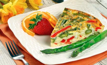 A vibrant slice of quiche with red peppers and asparagus, accompanied by fresh strawberries and orange slices, set on a decorative plate with floral accents in the background.