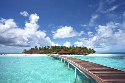 A tropical Maldives atoll with a wooden pier extending over clear turquoise sea toward lush islet tourist facilities under a bright blue sky with scattered clouds.