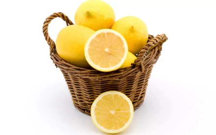 A wicker basket filled with fresh lemons, including a sliced lemon in front, set against a clean white background. A vibrant HD PC desktop wallpaper featuring fruit.