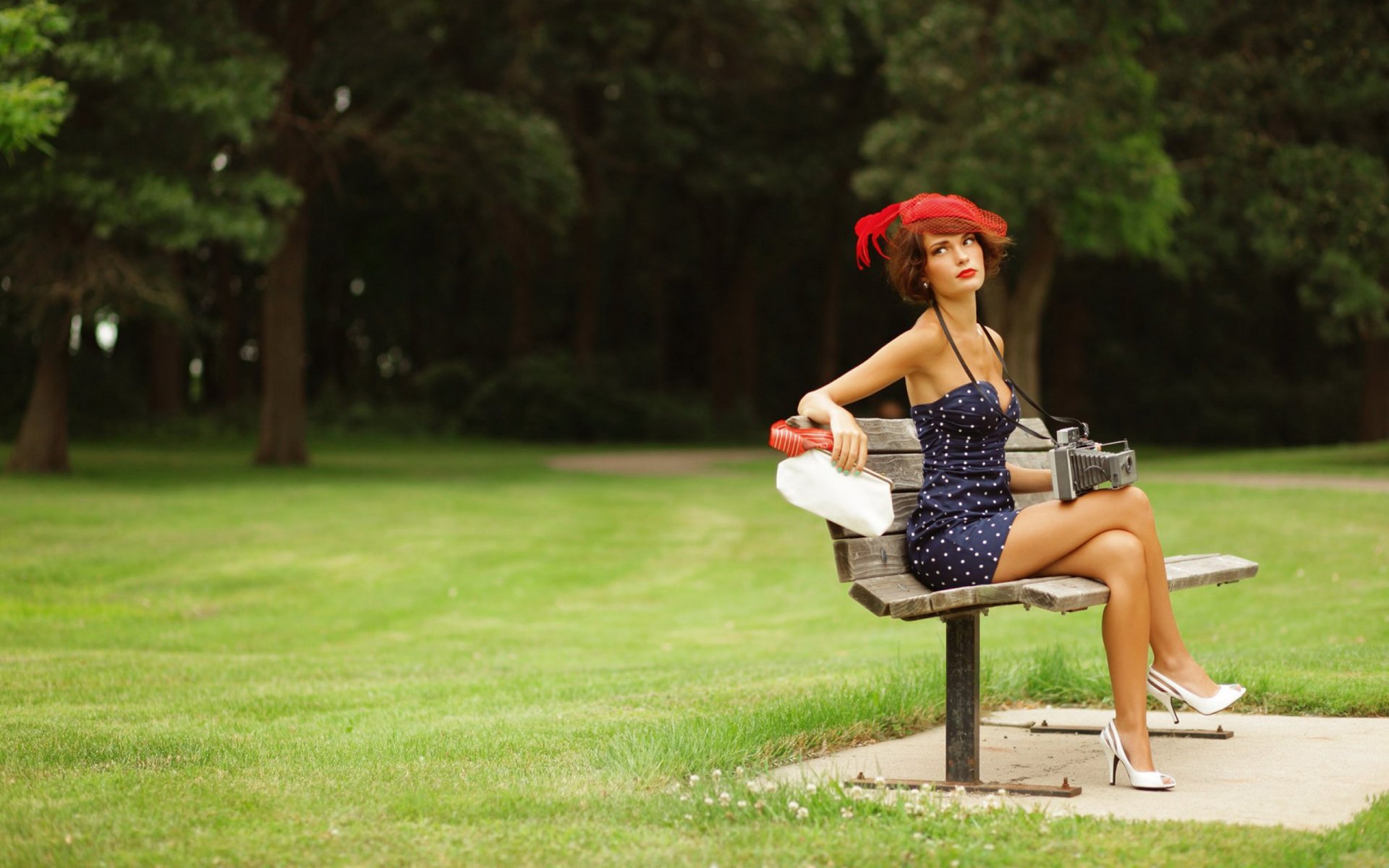 Woman model in a red hat and navy polka-dot dress seated on a park bench, holding a handbag — 2K Quad HD PC desktop wallpaper and background.