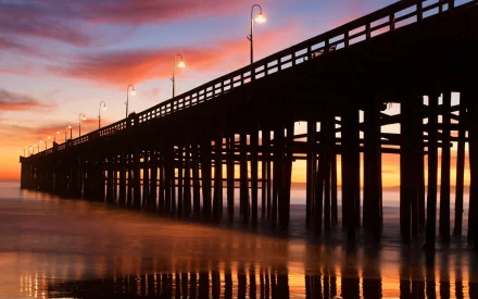 HD PC desktop wallpaper featuring a man-made pier extending over calm waters at sunset, with warm hues and glowing lampposts lining the structure.