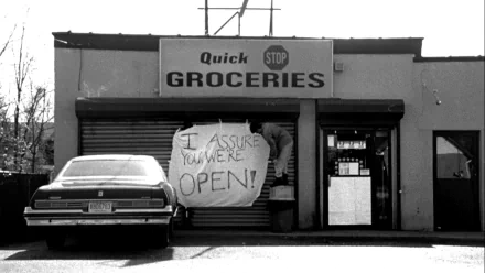 Black-and-white storefront from Clerks (movie): car parked outside Quick Groceries with a hand-lettered I ASSURE YOU WE'RE OPEN! sign — HD PC desktop wallpaper and background.
