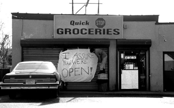 Black-and-white storefront from Clerks (movie): car parked outside Quick Groceries with a hand-lettered I ASSURE YOU WE'RE OPEN! sign — HD PC desktop wallpaper and background.