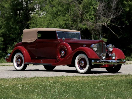 A vintage red Packard Eight convertible with a tan top parked on a grassy area, showcasing its classic design against a blurred natural backdrop.