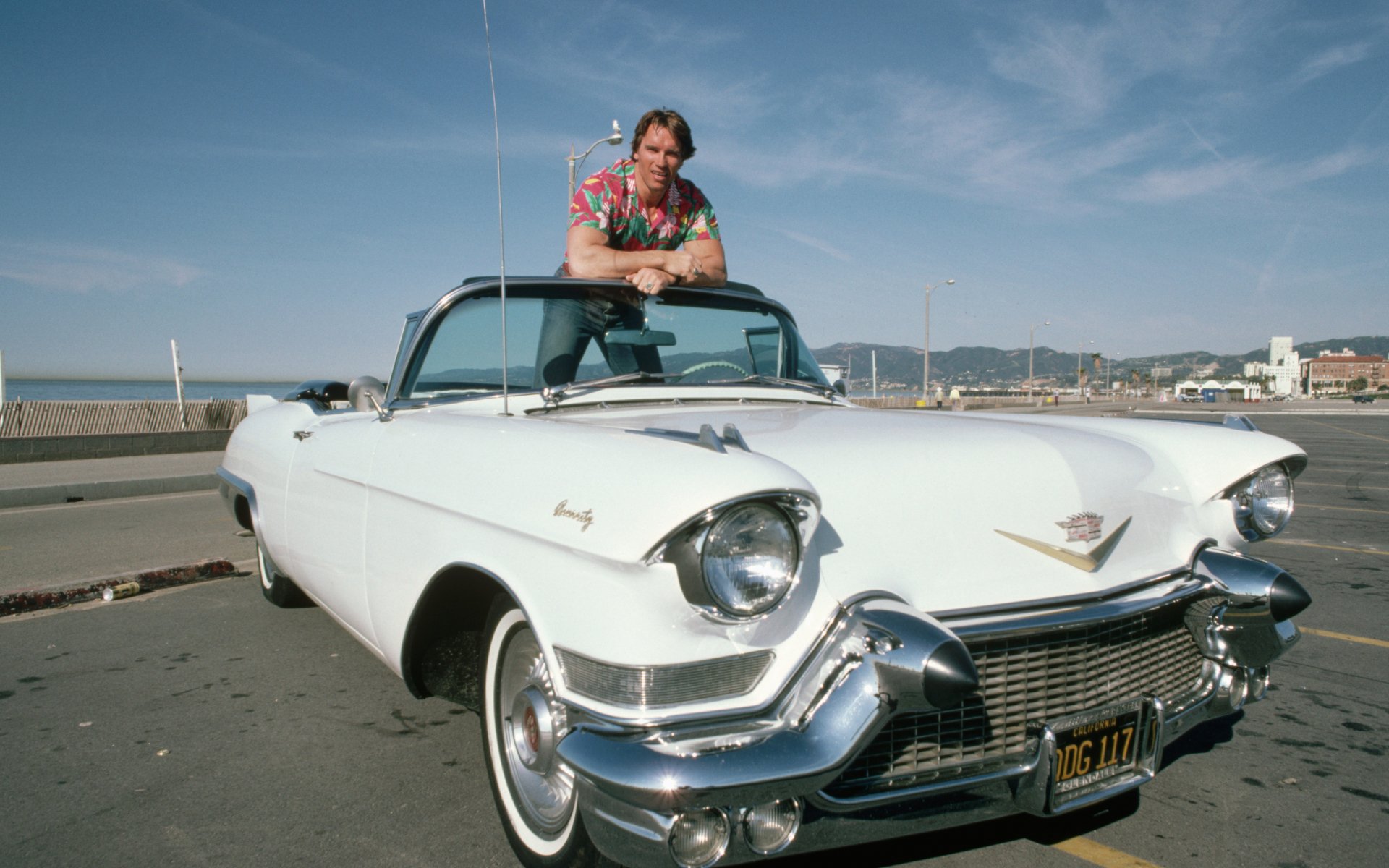 Arnold Schwarzenegger poses atop a classic white Cadillac convertible, set against a scenic backdrop, showcasing a vibrant and nostalgic vibe in this HD desktop wallpaper.