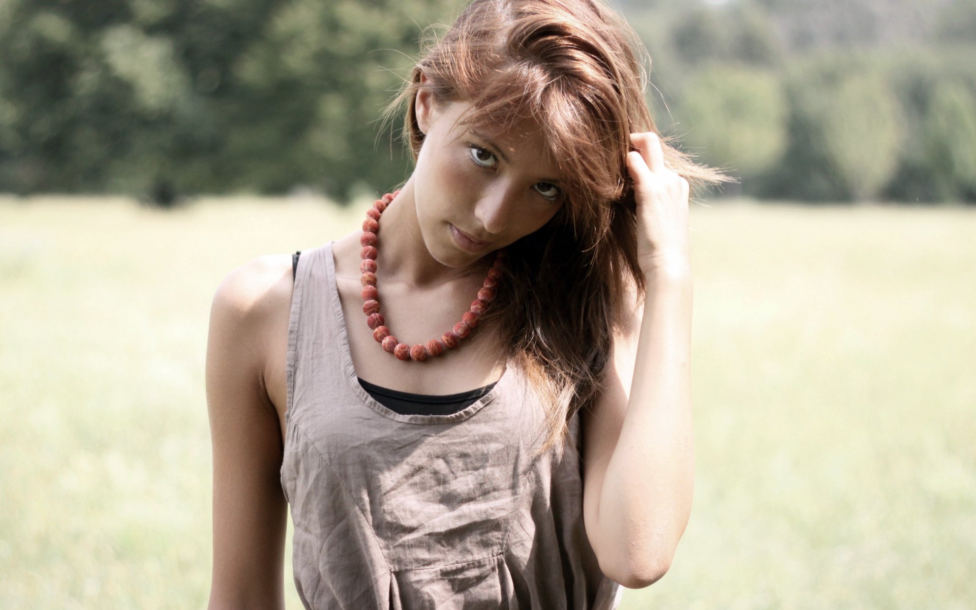 A woman stands in a field, gazing thoughtfully while running her fingers through her hair. She wears a simple outfit and a beaded necklace, conveying a serene mood. HD desktop wallpaper.
