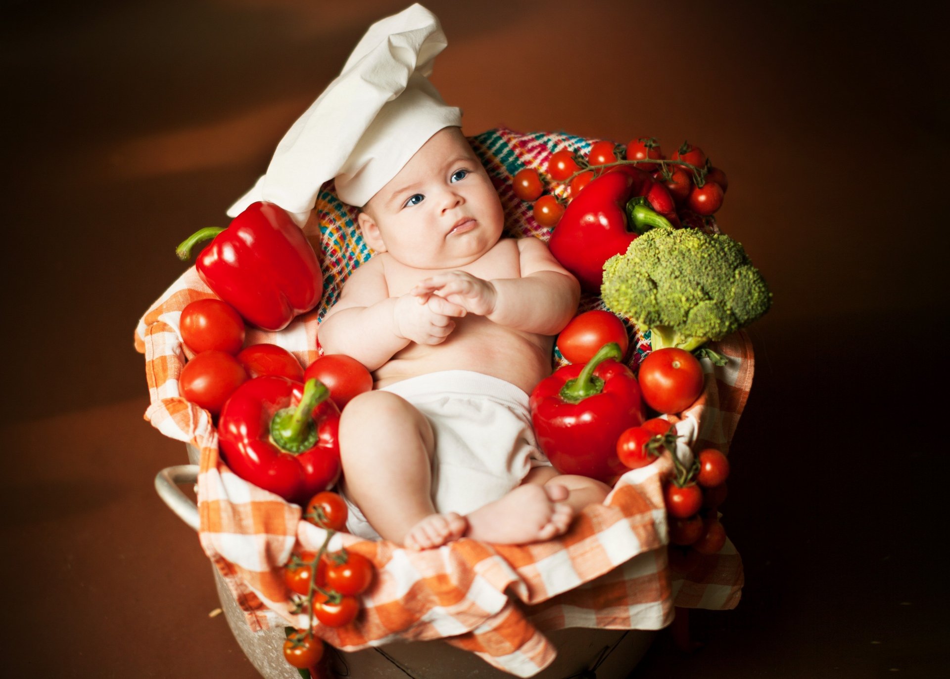 4K Ultra HD photography of a baby wearing a chef hat, surrounded by fresh vegetables like tomatoes, broccoli, and bell peppers on a checkered cloth.