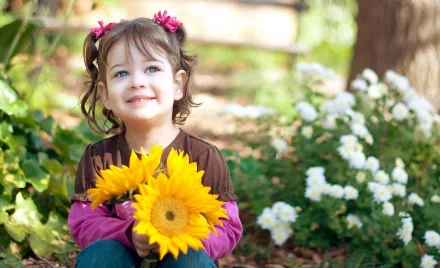 HD wallpaper of a child holding a bunch of bright yellow sunflowers, sitting in a garden surrounded by lush greenery and white flowers. The child is smiling and has pink flower hair accessories.