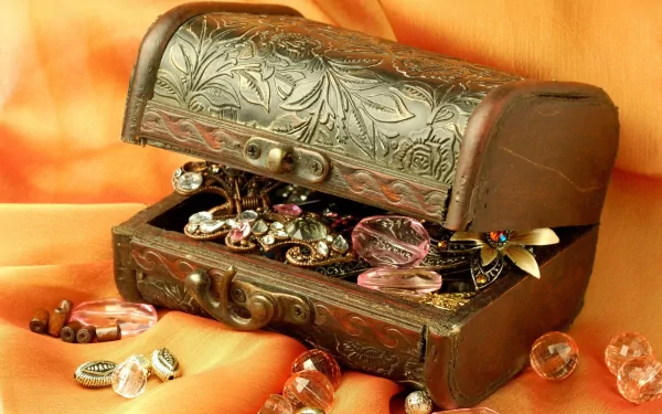 HD desktop wallpaper showing an ornate man-made jewelry chest overflowing with rings, bracelets, and decorative beads against a warm orange fabric background.