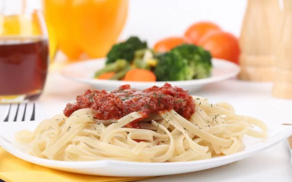 HD PC desktop wallpaper featuring a close-up of pasta topped with tomato sauce, with blurred vegetables and drinks in the background, emphasizing food and pasta.