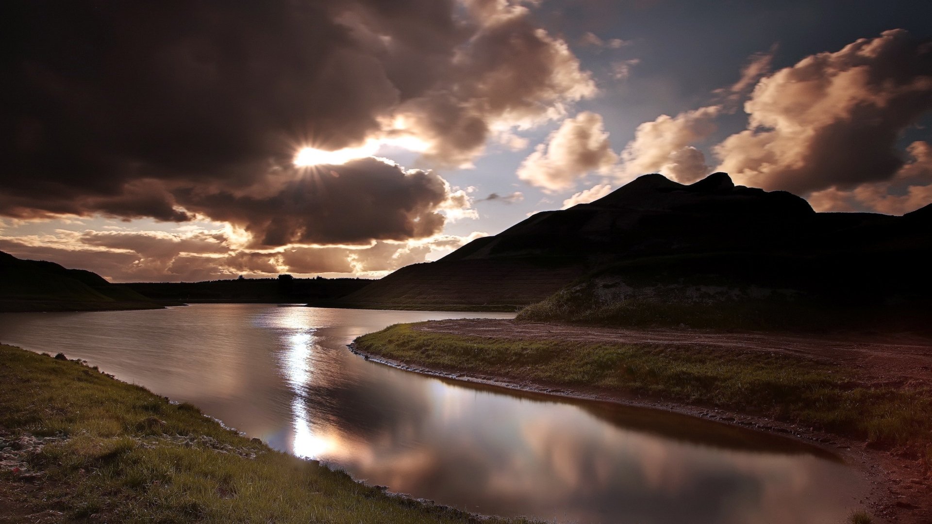 HD PC desktop wallpaper of a serene landscape featuring a river reflecting dramatic clouds and silhouetted hills at sunset.