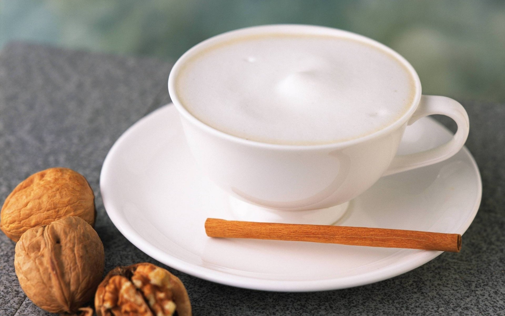 A soothing cup of frothy coffee accompanied by walnuts and a cinnamon stick, set against a blurred background, creating a serene and inviting atmosphere for a desktop wallpaper.