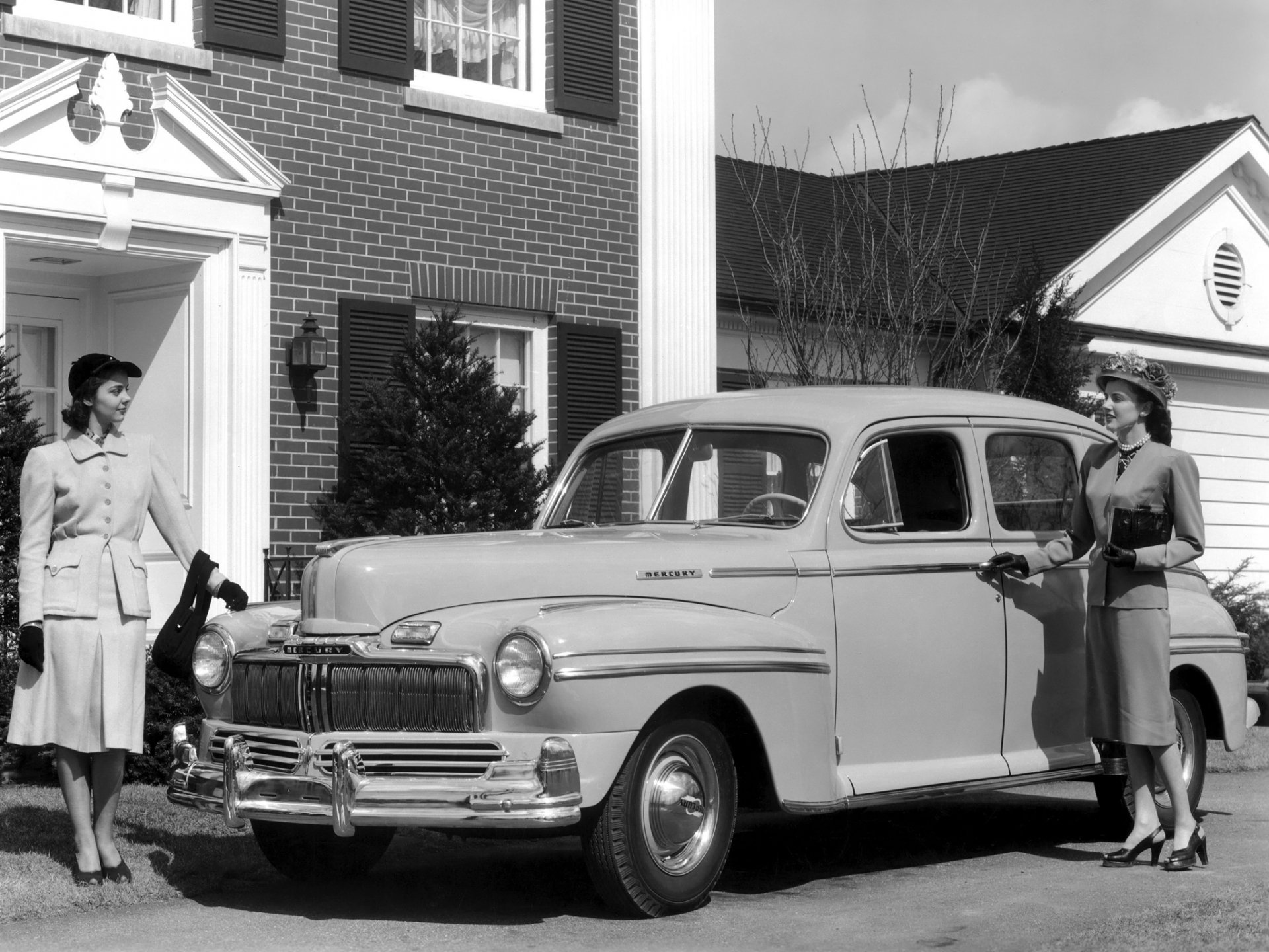 Black and white vintage photo of a 1947 Mercury Eight Town Sedan parked in front of a brick house, featuring two women in period attire.
