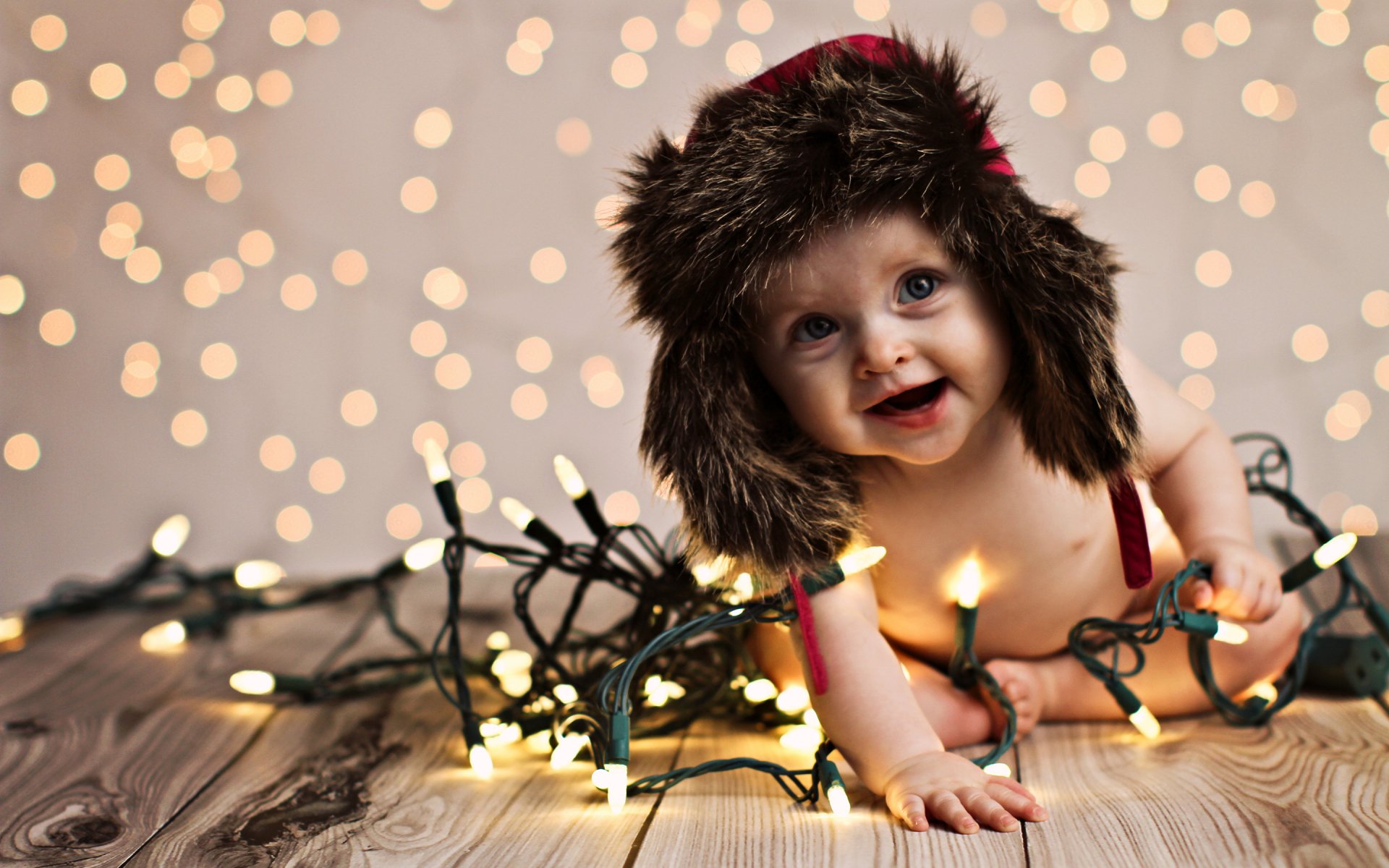 A joyful child wearing a furry hat crawls amid glowing Christmas lights against a bokeh backdrop, creating a heartwarming HD desktop wallpaper and background.