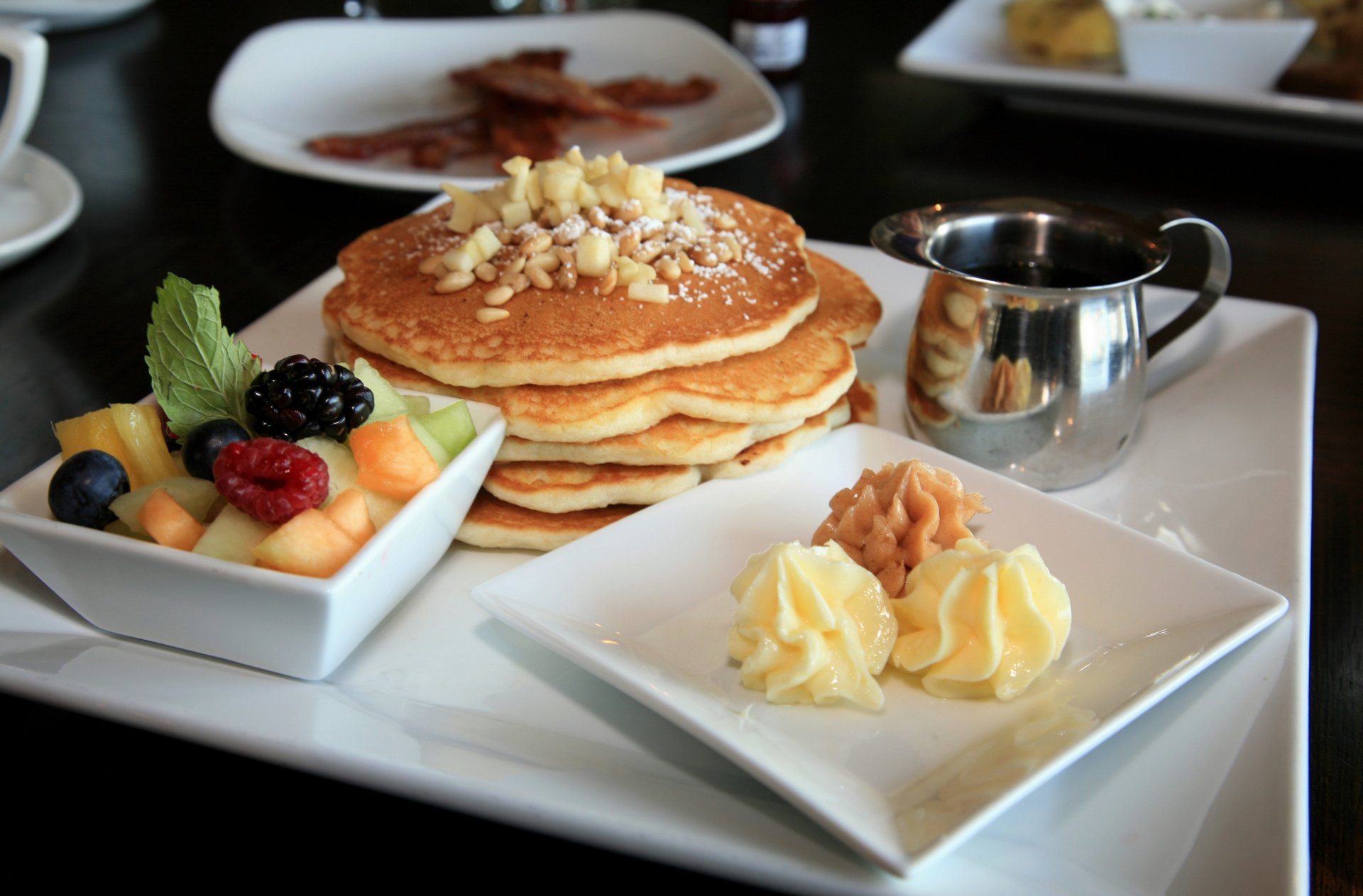 A stack of fluffy pancakes topped with nuts, accompanied by butter, fruit medley, and a small pot of syrup, captured in a vibrant 4K Ultra HD background.