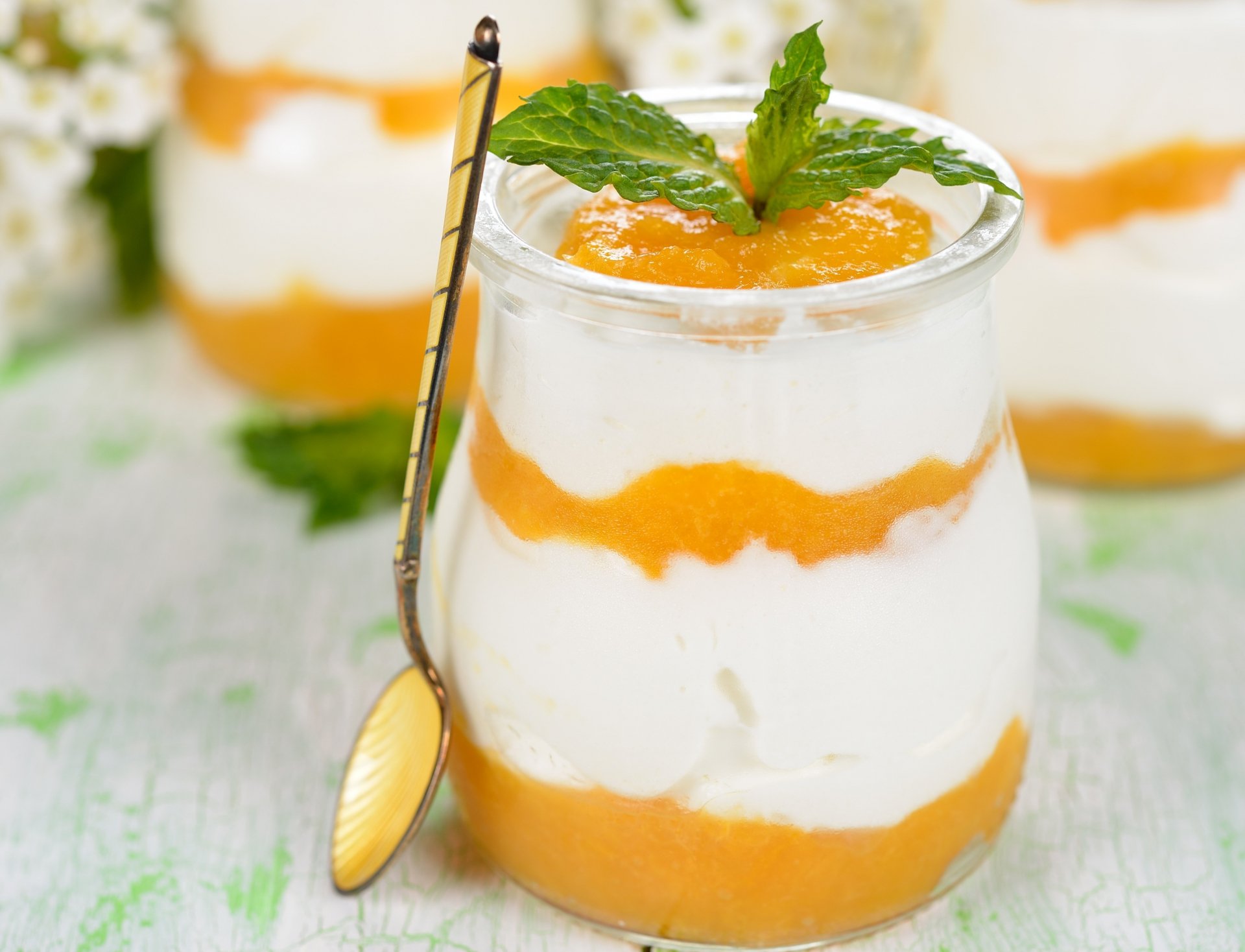 A close-up 4K Ultra HD dessert wallpaper showing a glass filled with layered creamy white yogurt and bright orange fruit puree, garnished with fresh mint leaves and a spoon.